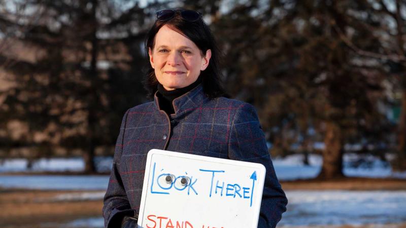 Megan Pickett poses for a photo on Main Hall Green while holding a small white board that reads Stand Here and Look There, with an arrow pointing to the sky.