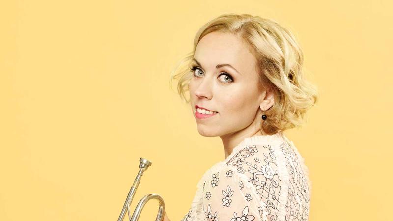 Trumpet player Tine Thing Helseth holds her trumpet looking back at the camera over her left shoulder against a yellow background.