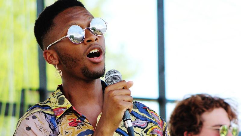 Student sings on outdoor stage. He is wearing reflective sunglasses and a colorful shirt