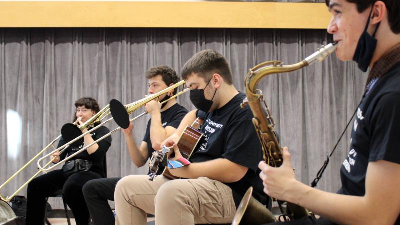 Four students with various instruments, such as saxophone and trumpet, sit in a row in a large room.
