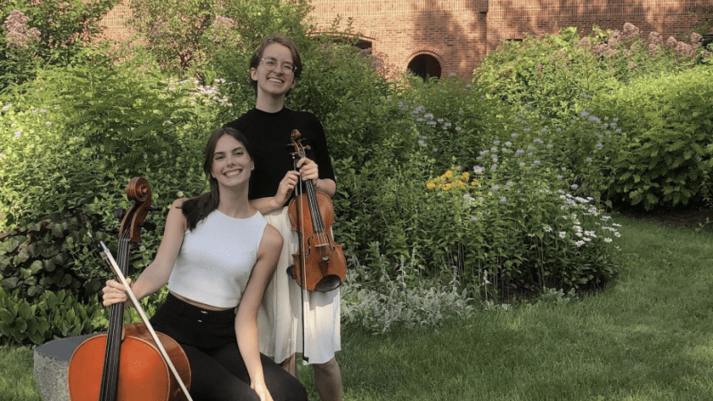 Two students hold a violin and cello in a garden.