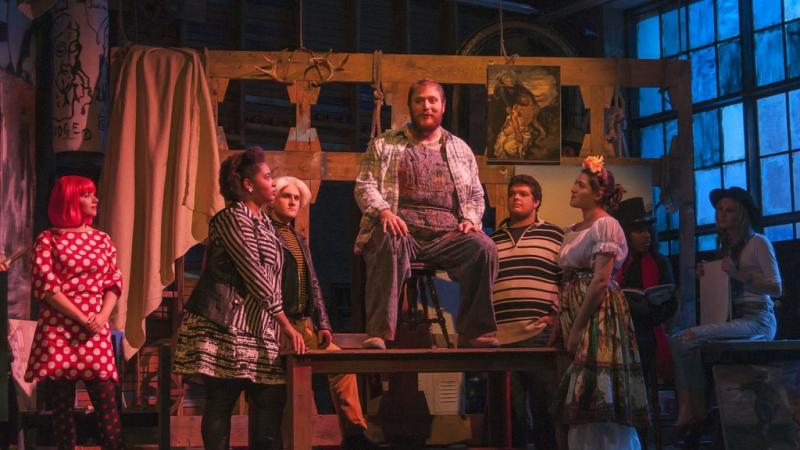 Production photo of the musical, Godspell: man sitting on chair on top of table while 6 others stand around the table and watch him.