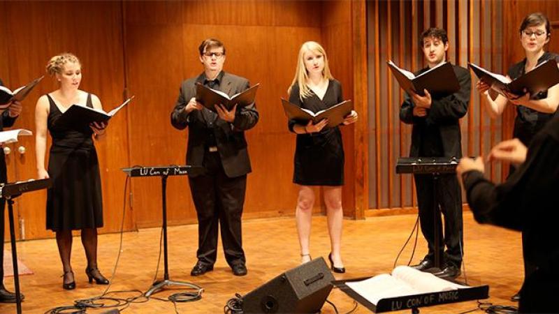 Seven singers in black stand in semi-circle with music stands in front of them on stage.