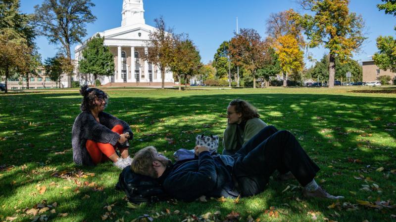 Three students sitting on Main Hall Green on sunny day with Memorial Chapel in background.