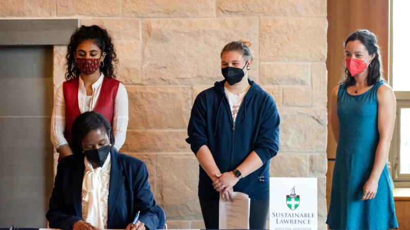 President Laurie Carter signs the Climate Commitment at a table while Adya Kadambari, Em Gajewski and Relena Ribbons stand behind her.