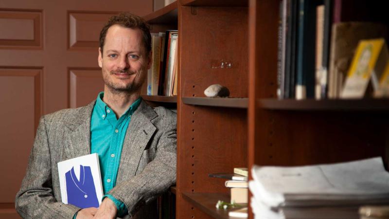 Austin Segrest holds his newly published book while leaning against a book shelf in his office.