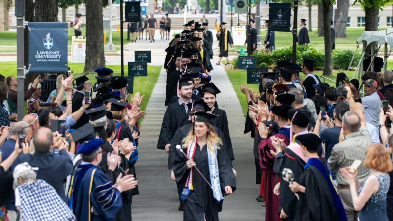 Graduates dressed in cap and gown process between rows of faculty dressed in regalia.