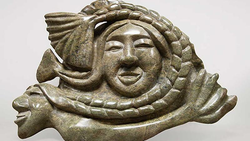 Green stone carving of a woman's face surrounded by a fish tail braid, supported by a prone figure of a man with a fish tail.