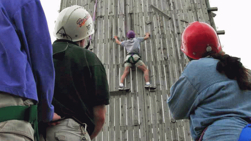 Student climbing outdoor rock wall while three others watch from below.