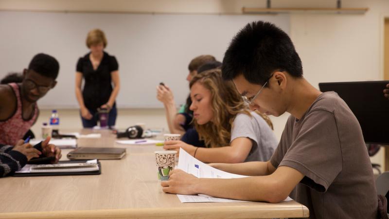 Summer Institute students reading a Workshop Handout in a Briggs Hall Classroom. Kate Zoromski, the Workshop host is standing at the end of the table.