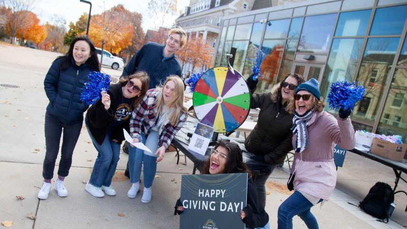 Students and staff hold a Happy Giving Day sign in front of the Spin the Wheel game in front of Warch Campus Center.