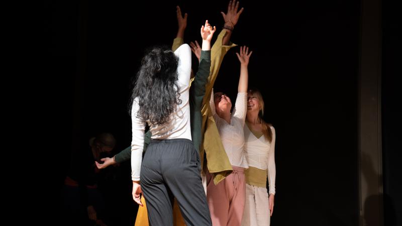 Dancers stand in a clump with arms reaching upwards in a dark room.