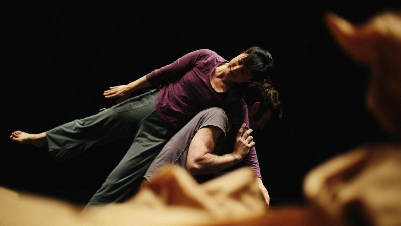 Dance Intensive one dancer is crouched down and another is leaning over the dancer with side on crouched dancers back and arm over shoulder