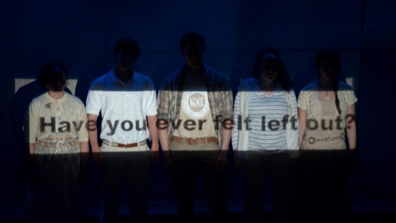 Scene from performance "Show Your Face(s): A Masque" where students stand against background and the question "Have you ever felt left out?" is projected across them.