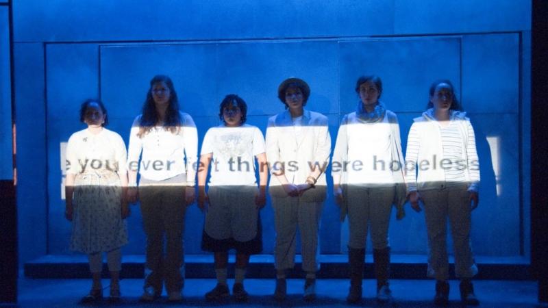 Scene from performance of "Show Your Face(s): A Masque" where students stand in a row and the question "Have you ever felt things were hopeless?" is projected across them.
