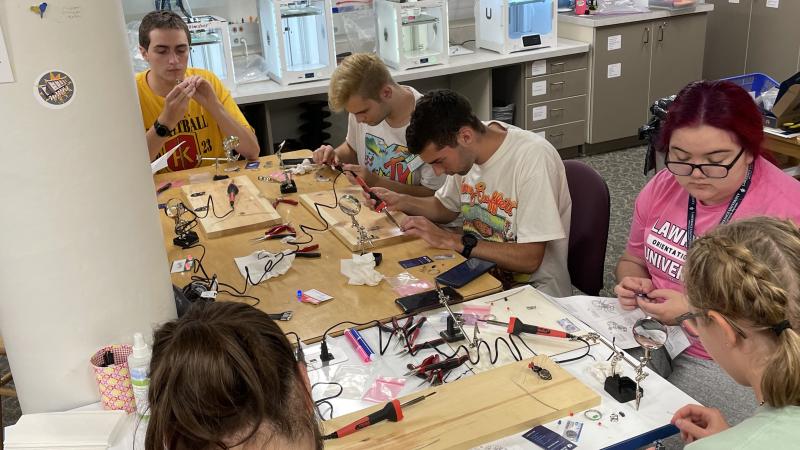 Students sitting around a table practicing soldering in the makerspace with 3D printers printing in the background.