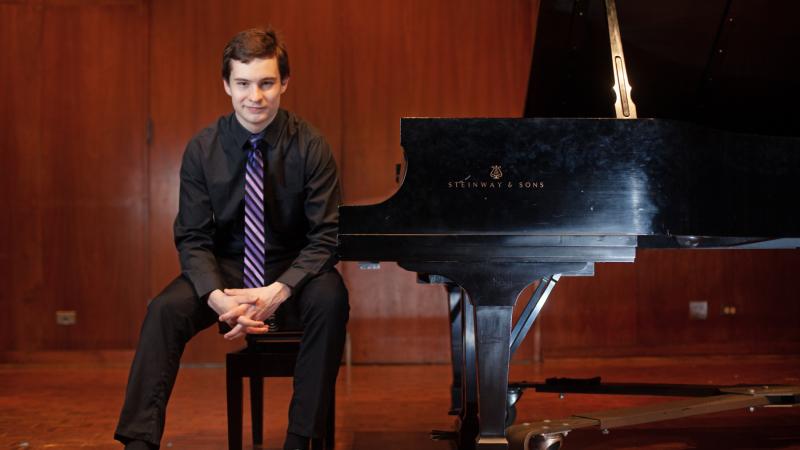 Jonathan Bass sits on a piano bench next to a piano.