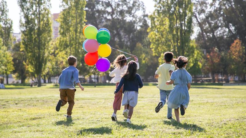 a group of children running away from the camera, holding a bunch of balloons