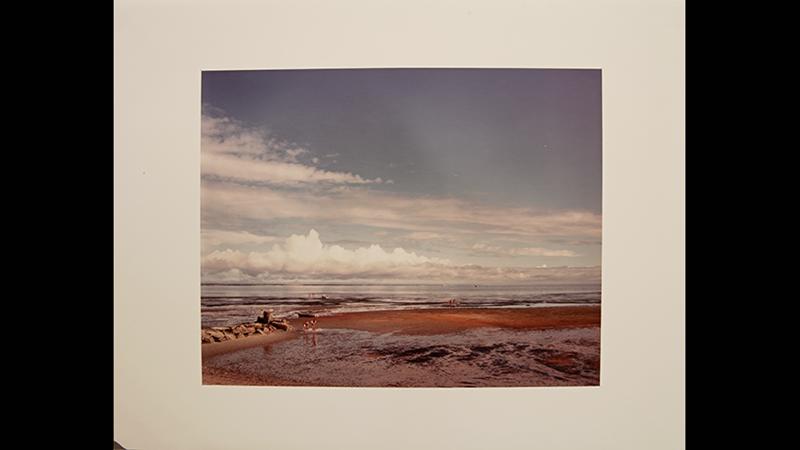 A color photograph of a beach and a sand bar, with another inner body of water. Groups of people walk along the beach. The frame is dominated by a blue sky with clouds.