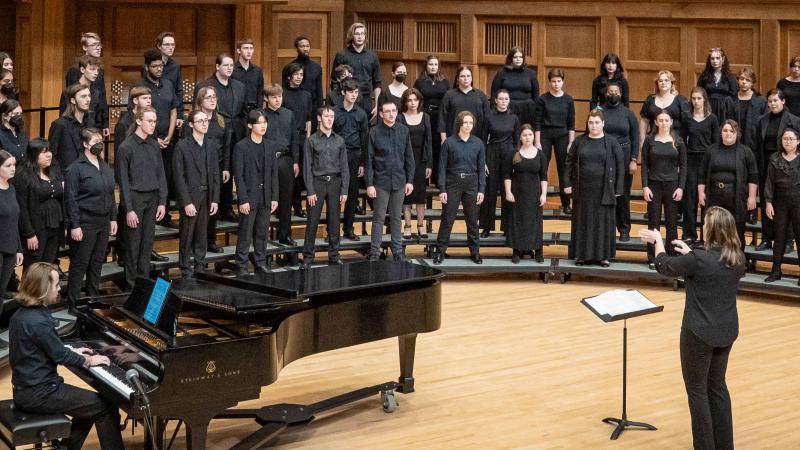 A choir dressed in concert black accompanied by a pianist sings on risers in the chapel while their director gestures in front of the group