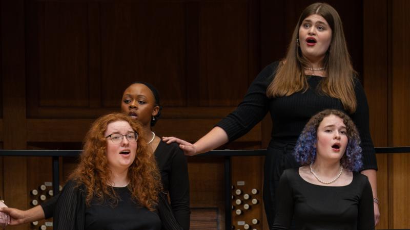 Four women in concert black sing on stage, two holding hands and one touching her neighbor's shoulder