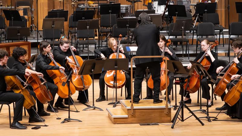 A cello ensemble performing on stage, led by a conductor.