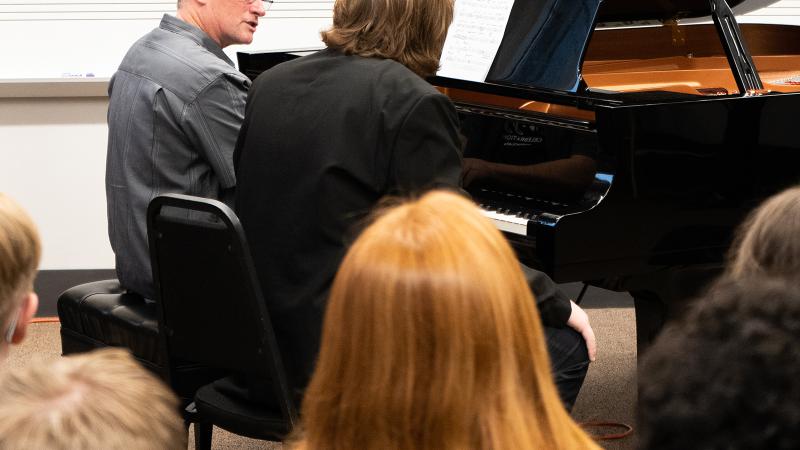 A small group of people observing a professor teaching a student at a piano.
