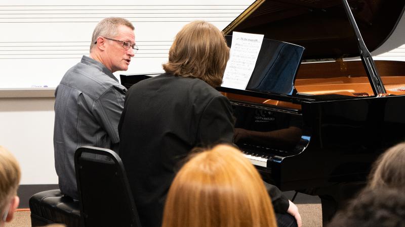 A small group of people observing a professor teaching a student at a piano.