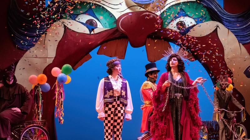 A small ensemble of costumed performers on a stage with a gigantic clown face overhead.
