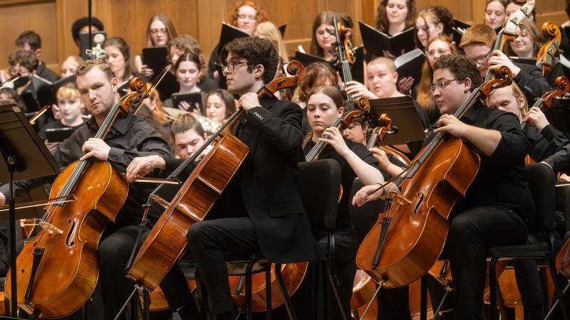A group of musicians playing cello, with choral singers in the background.