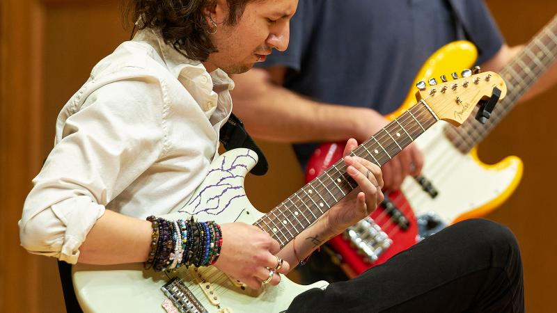 A student focused on playing electric guitar, with another student playing electric bass in the background.