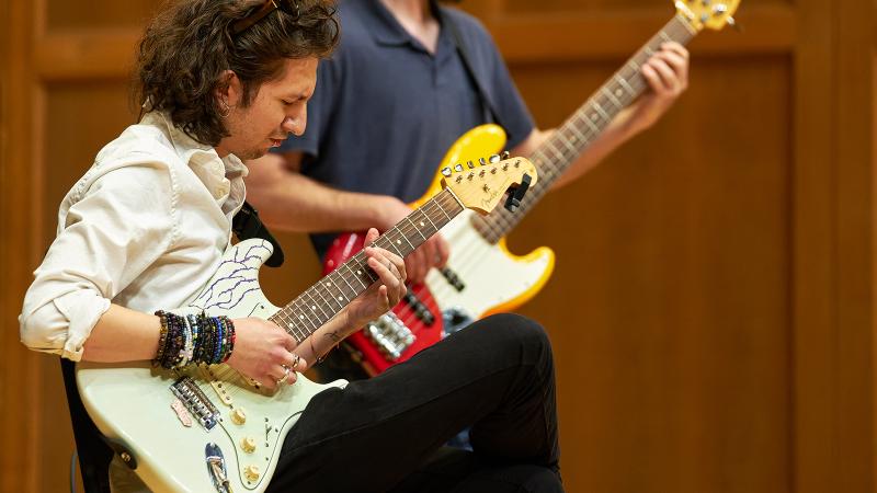 A student focused on playing electric guitar, with another student playing electric bass in the background.