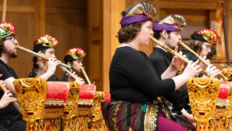 An ensemble of costumed musicians playing bamboo flutes while seated next to gold and red metallophone instruments.