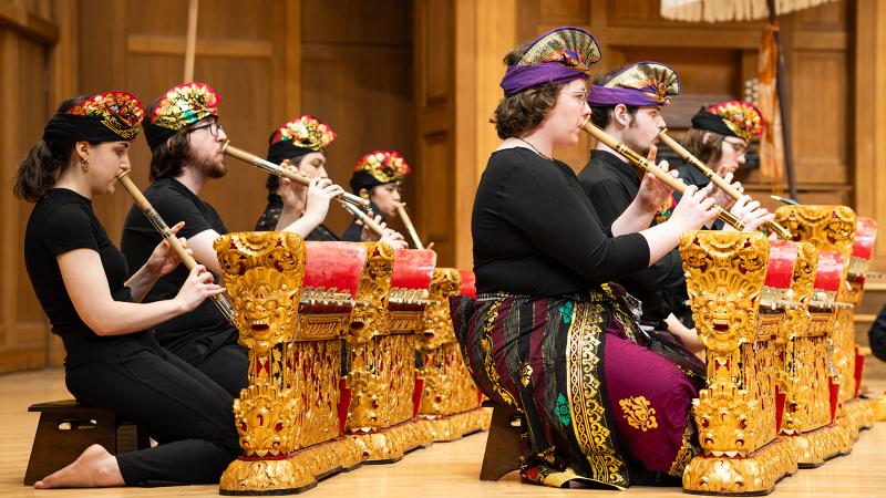 An ensemble of costumed musicians playing bamboo flutes while seated next to gold and red metallophone instruments.