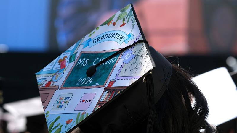 A graduation cap decorated with a scene of a K-12 classroom.