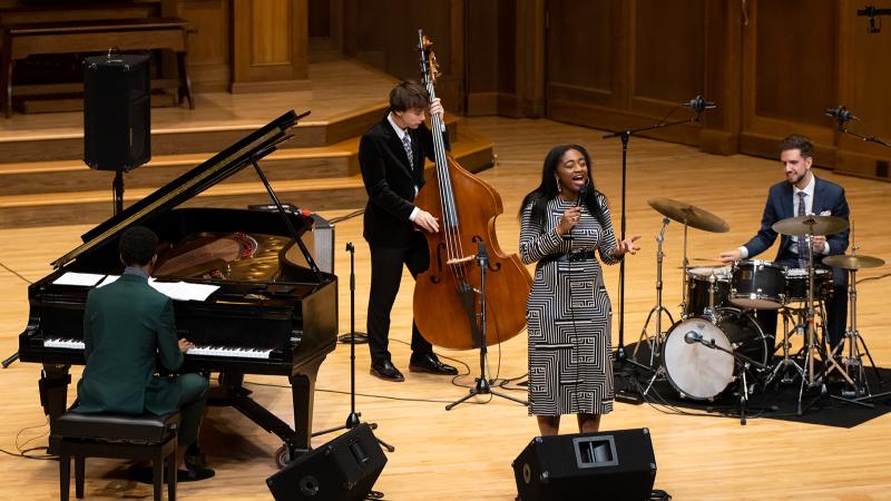 A jazz quartet of vocals, piano, bass, and drum set.