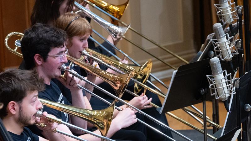 A small group of musicians playing trombones into a row of microphones.