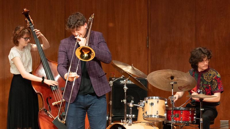 Three jazz musicians playing upright bass, trombone, and drum set in performance.