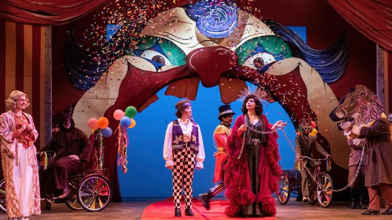 A small ensemble of costumed performers on a stage with a gigantic clown face overhead.