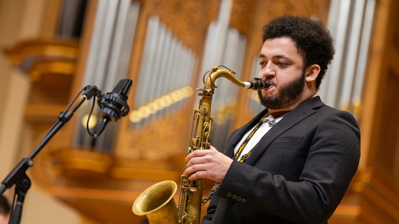 A musician playing tenor saxophone, with a pipe organ in the background.