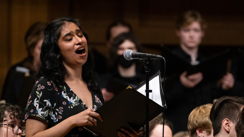 A vocalist singing a solo in performance, with an orchestra and chorus in the background.
