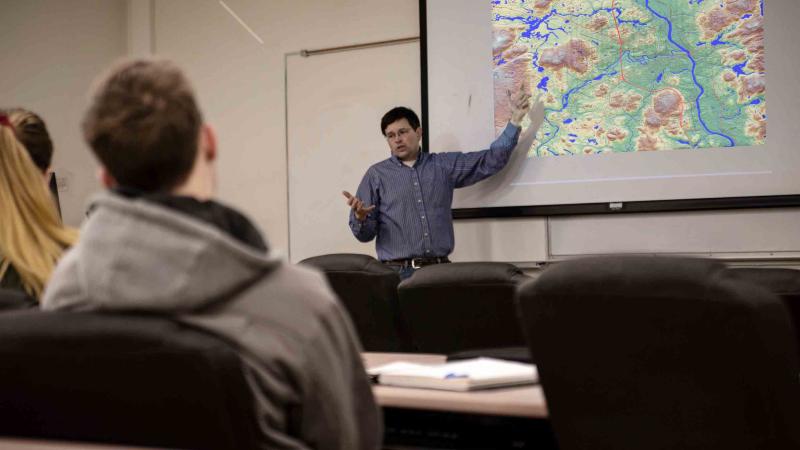 A professor pointing at a screen projection of a topographical map with students looking on from the classroom