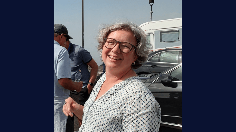 Photograph showing a person with gray hair wearing a white patterned shirt standing outdoors near parked cars and a trailer. 