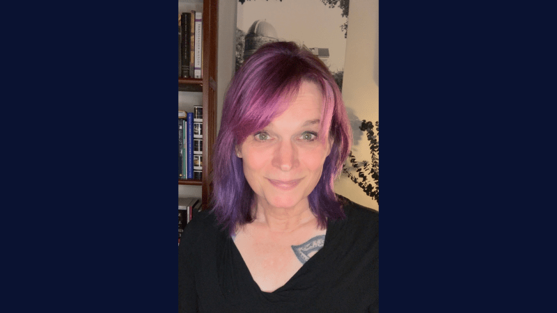 Photograph of a person with shoulder-length purple hair wearing a black top, standing indoors near a bookshelf and a wall with framed art. Visible tattoo on upper chest and soft lighting highlighting the side of the face and hair.