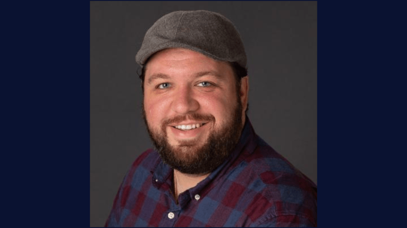 A man with a beard, wearing a gray cap and a red-and-blue plaid shirt, smiles at the camera against a dark background.