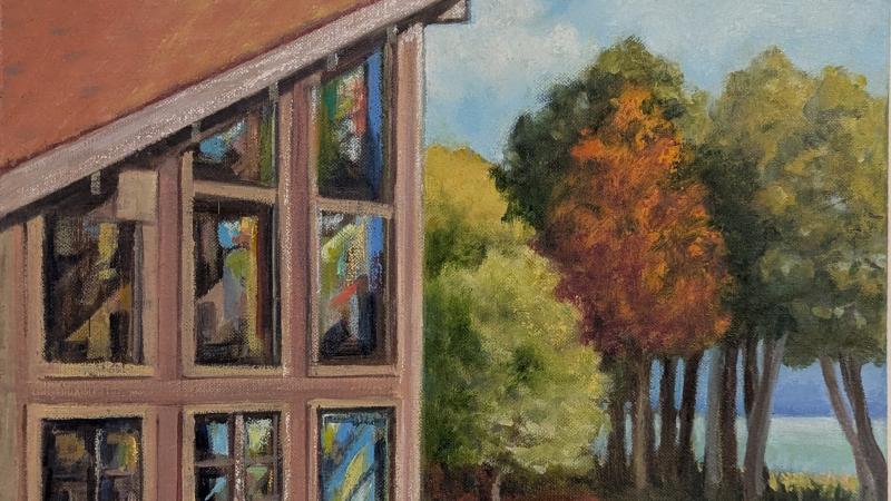 A view of Bjorklunden's 'Great Room' from the outside. The image points north and depicts the iconic windowed corner of the room in the foreground. Behind it sit autumn trees with leaves of oange, green, and gold. And behind them sit white cumulus clouds in a blue sky, suspended above the seemingly endless water of Lake Michigan.