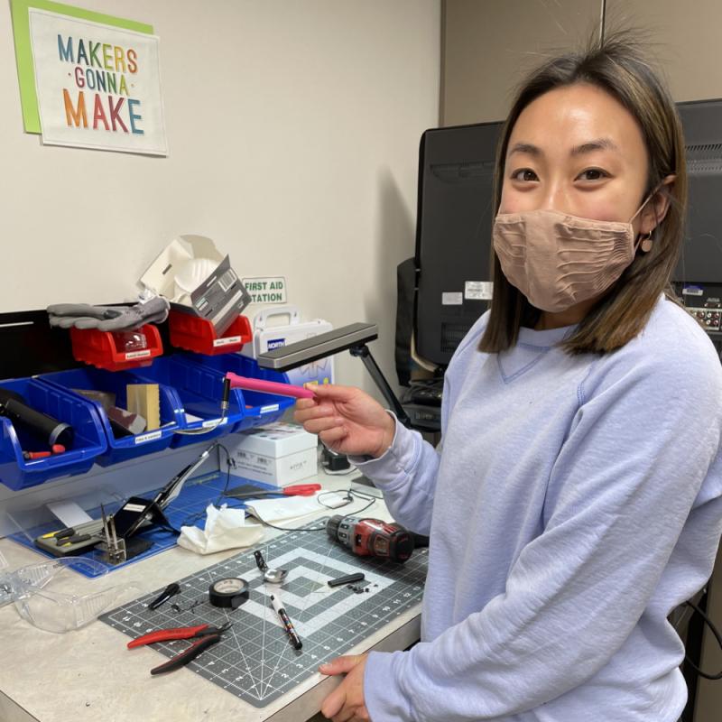 Woman holding a custom 3D printed accessible swivel spoon she made near a workspace full of tools.