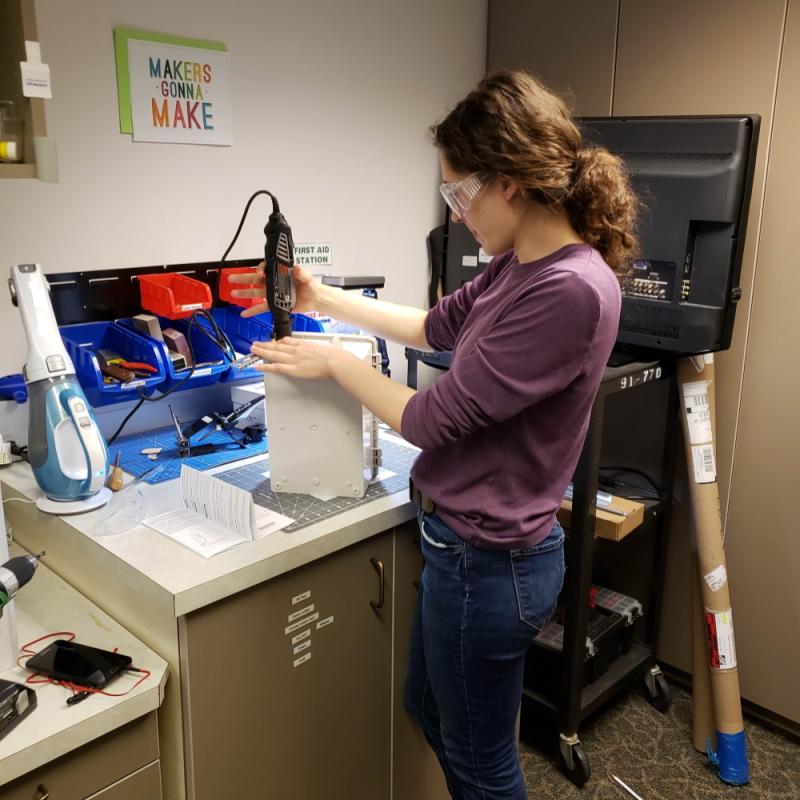 Woman standing at a workspace in the makerspace using a Dremel sander to smooth a hole in an object she 3D printed.