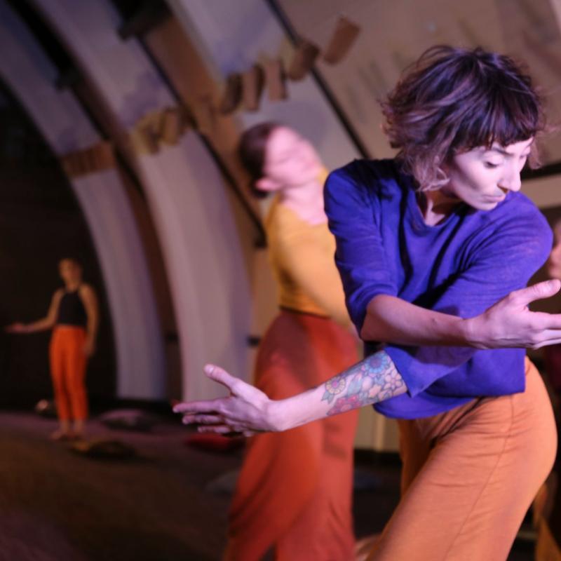 A light skinned woman with short brown hair and bangs twists upper body and crosses arms. Her head is turned, looking over her left shoulder. She is dressed in a purple top and orange gold pants. the photo is lit from the right corner, stage lights gently illuminating her body.