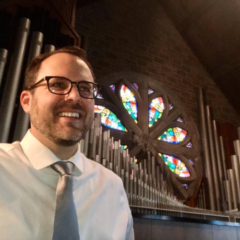 Man with glasses in front of organ pipes and stained glass window.